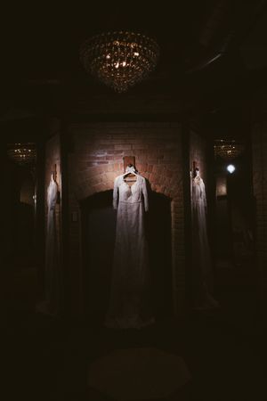Elegant white wedding dress displayed on hanger in dimly lit room with crystal chandelier and brick wall backdrop.