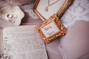 Elegant wedding flat lay with gold frame earrings, open journal, pearl necklace, and rings on soft pink background.