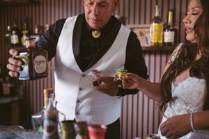 Bartender in white vest pours whiskey bottle while bride in strapless gown holds colorful shot glasses.