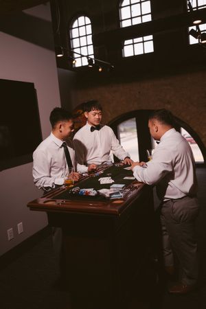 Three men in white shirts play poker at a wooden table in a dimly lit room with arched windows.