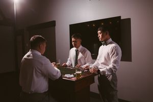 Three men in white dress shirts and ties gather around a small table in a dimly lit room for a groomsmen meeting.