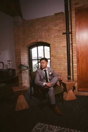 Groom in gray suit sitting in chair before wedding ceremony in rustic brick venue.