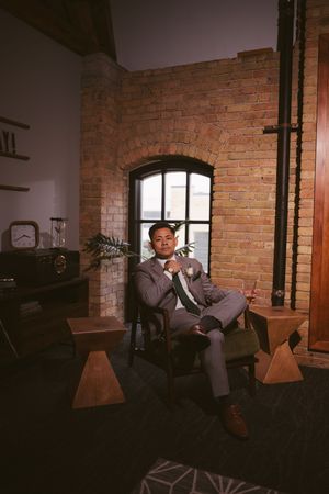 Groom in gray suit posing in chair with boutonniere in elegant brick-walled venue.