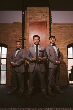 Three groomsmen in gray suits and bow ties pose formally in front of a brick wall and wooden door.