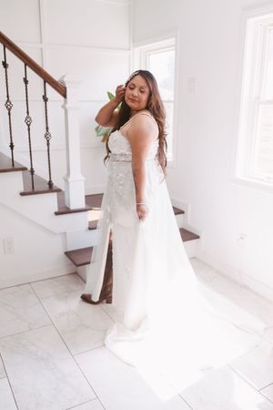 Bride in elegant white lace wedding gown with slit stands by staircase in bright white interior.