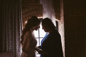 Two women share an intimate moment in silhouette by a window, one in a wedding dress, in a warmly lit brick room.