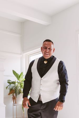 Man in white vest and black shirt smiles confidently indoors with plant and window in background.
