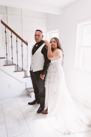 Bride and groom pose elegantly by a staircase in a bright white interior, she in a lace gown, he in a black tuxedo.
