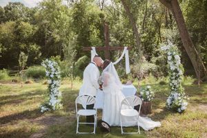 Bride and groom share first kiss at outdoor wedding ceremony with white floral arch and lush green trees.