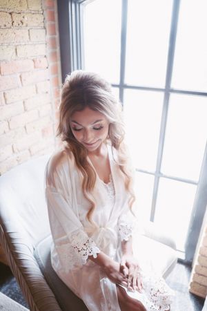 Elegant bride in white lace robe sitting by window with soft natural light, showcasing bridal morning preparation.