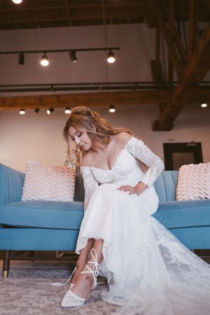 Bride in lace off-shoulder wedding gown sitting on blue sofa, tying white heels in elegant indoor venue.