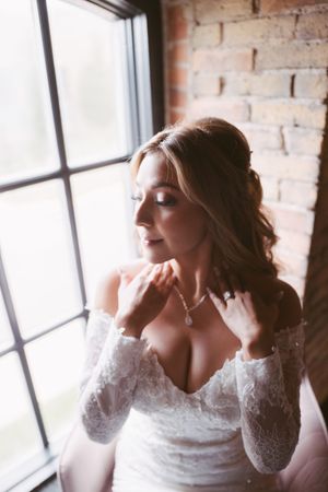 Bride in lace off-shoulder gown fastening pearl necklace by sunlit window in brick venue.