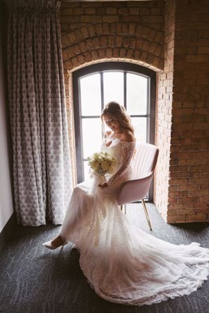 Bride in lace gown holding bouquet seated by arched window with brick wall backdrop.