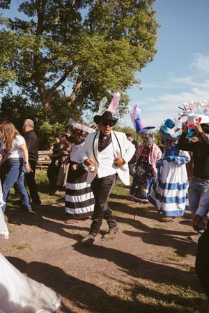 People in colorful traditional costumes dance outdoors near trees at a festive cultural celebration.