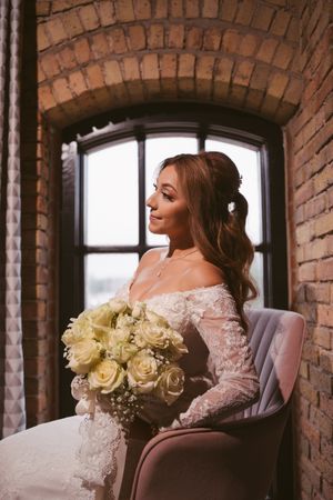 Bride in off-shoulder lace gown holding white rose bouquet seated by arched brick window.