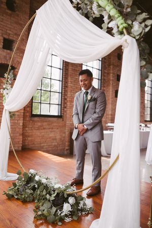 Smiling groom in gray suit standing at floral wedding arch with white draping in brick venue.