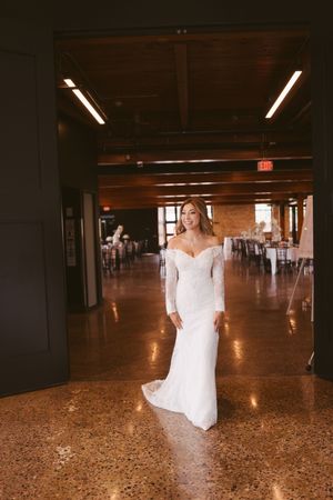 Bride in fitted off-shoulder lace wedding gown standing in elegant indoor venue hall.