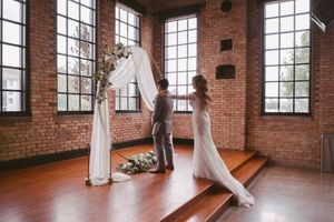 Bride and groom arranging a floral wedding arch with white draping in an industrial brick venue.