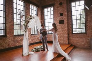 Bride and groom exchanging vows under a white floral arch in a rustic brick-walled wedding venue.