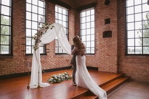 Bride in flowing gown adjusting white draped floral arch on wooden stage in brick ceremony hall.