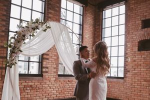 Bride and groom share a romantic moment under a floral arch with white draping in a rustic brick venue.