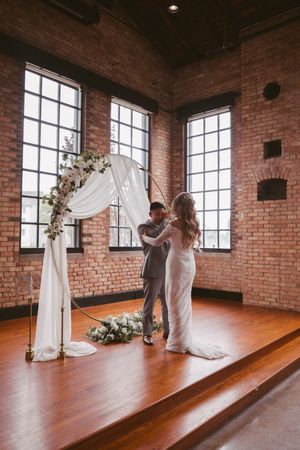 Bride and groom exchange vows under a floral hoop arch with white draping in a rustic brick wedding venue.