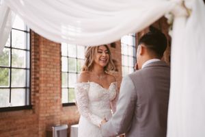 Bride in lace off-shoulder gown laughing joyfully with groom during wedding ceremony in brick venue.
