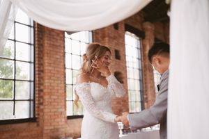 Emotional bride wiping tears holding grooms hands during heartfelt wedding ceremony in rustic brick venue.