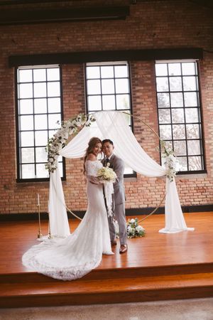 Bride and groom embracing under circular floral arch with white draping in brick venue.