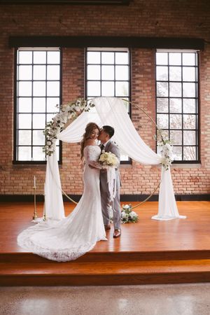 Bride and groom share a kiss under a circular floral arch with white draping on a wood stage in a brick venue.