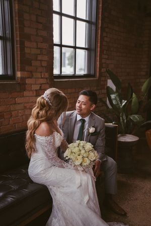 Bride and groom sitting on a leather sofa holding white roses bouquet in a brick-walled wedding venue.
