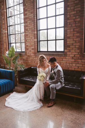 Bride and groom sitting on black leather sofa in front of brick wall and large windows, bride holding white bouquet.
