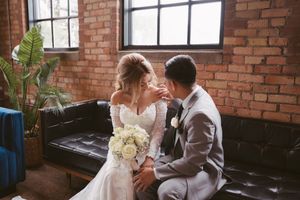 Bride and groom share an intimate moment on a black leather sofa in a rustic brick-walled wedding venue.