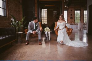 Bride in white lace gown and groom in gray suit relaxing in leather chairs at industrial wedding venue.