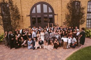 Large wedding party group photo outside brick venue with arched doors and landscaping.