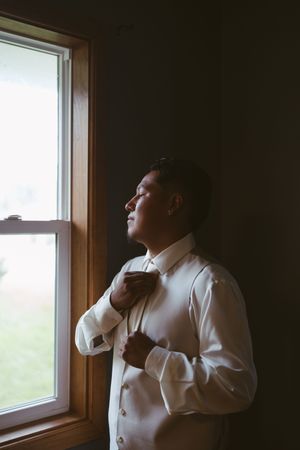 Groom adjusting bow tie by window in white dress shirt before wedding ceremony.