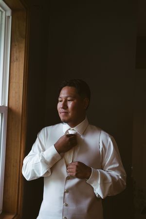 Groom in white dress shirt and vest adjusting bow tie near window in soft natural light.