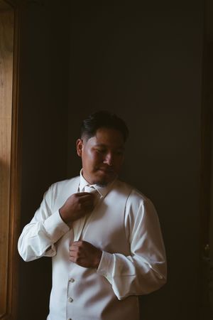 A groom in a white dress shirt adjusts his tie while getting ready, standing in soft window light.