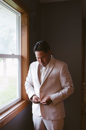 Groom in a beige suit buttoning his jacket by a window on his wedding day.