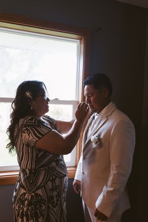 A woman in a patterned dress adjusts a groom's tie near a window before a wedding ceremony.
