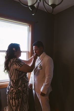 A woman in a sequined dress adjusts a groom's boutonniere as he stands in a white suit near a sunlit window.