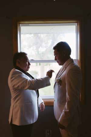 Two men in white tuxedo jackets share a moment by a window, one adjusting the other's boutonniere.