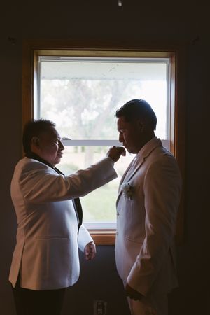 Two grooms in white tuxedos share a tender moment by a window as one adjusts the others boutonniere.