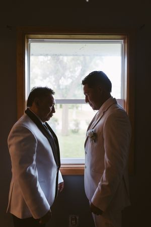 Two grooms in matching white tuxedos share an intimate moment standing face to face by a sunlit window.