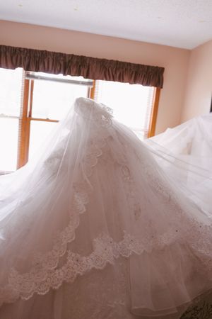 Elegant white lace wedding dress displayed in a sunlit room with brown curtain valances.
