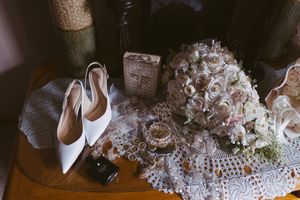 White bridal heels beside a pink rose bouquet, lace doily, perfume bottle, and vintage decor on a wooden table.