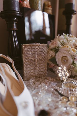 Wedding details including bridal shoes, rosary, bouquet, and ornate prayer book on a lace table.