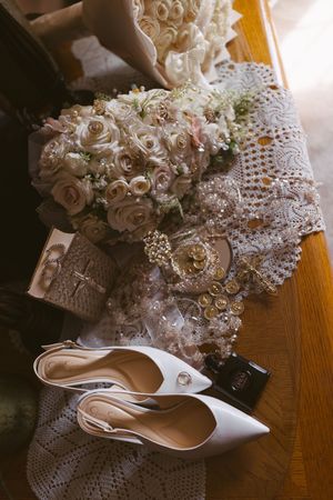 Elegant bridal accessories including white pointed heels, pearl bouquet, lace gloves, and gold jewelry arranged on a wooden surface.