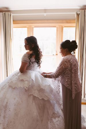 A bride in an ornate white lace ball gown being helped by a woman in a beaded dress near a bright window.