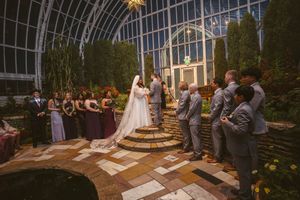 Elegant wedding ceremony in a stunning greenhouse venue with bridal party in burgundy and groomsmen in gray suits.